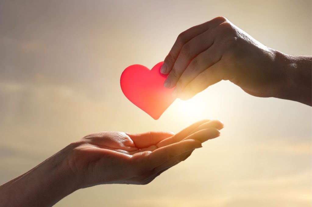 A hand extended waiting to receive a heart from another hand. Sunset sky in the background.