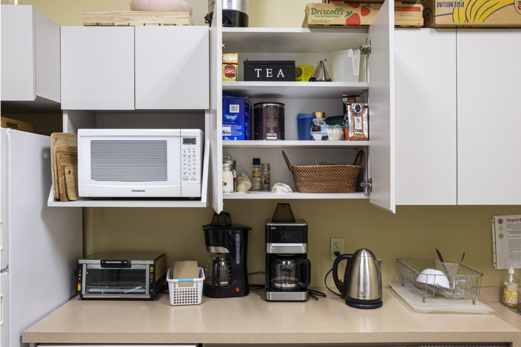 GCF office kitchen counter showing kettle, microwave, coffee pots, toaster over, cutting boards and a cabinet open to show an assortment of teas and coffee.
