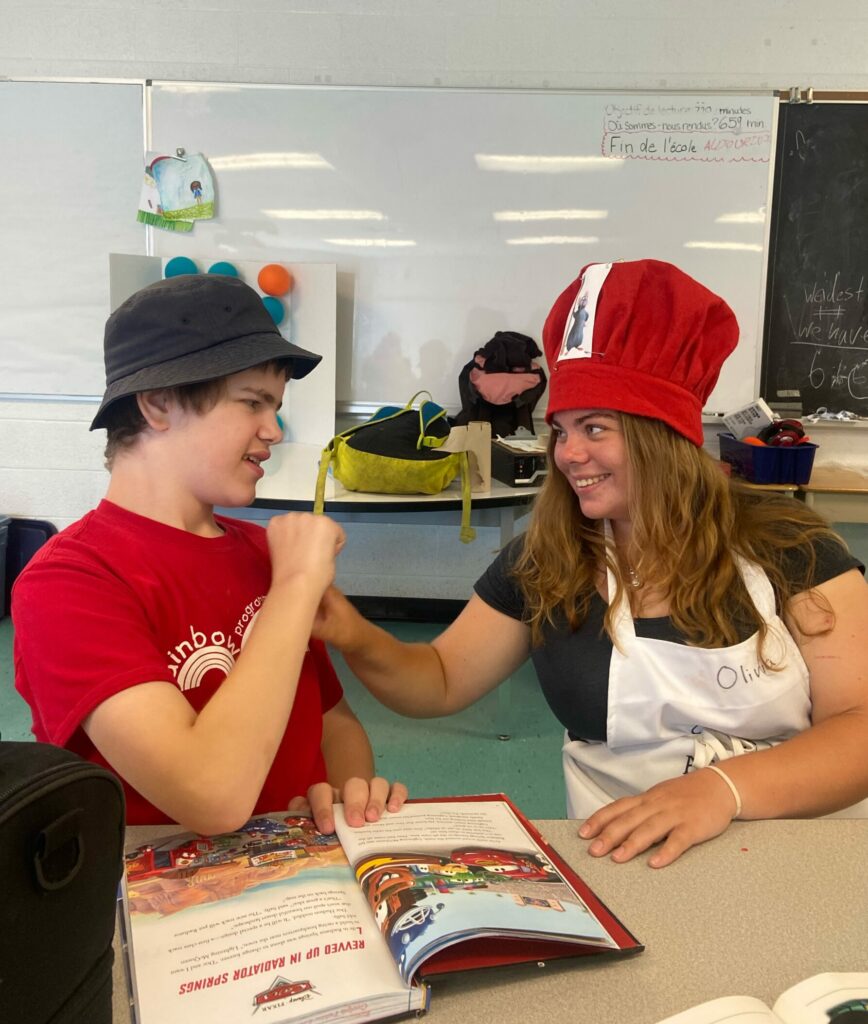 Kids to Camp 2022 Rainbow Camp For Children Photo of camper and counselor reading together wearing hats in a classroom