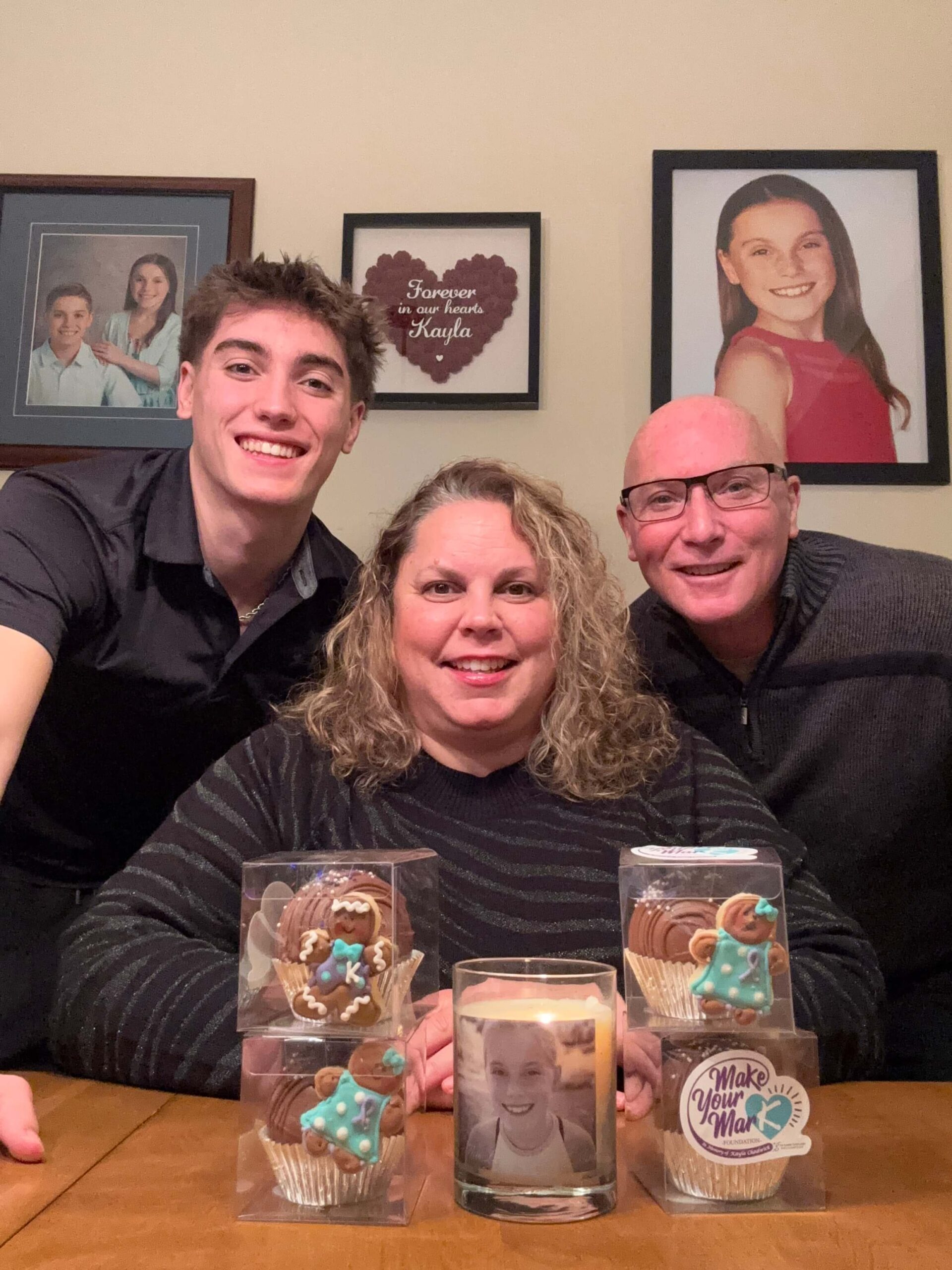 Image of Kayla's brother, mother and father at a table with pictures of Kayla on the wall behind them and examples of the hot chocolate bombs they sell as fundraisers on the table in front of them