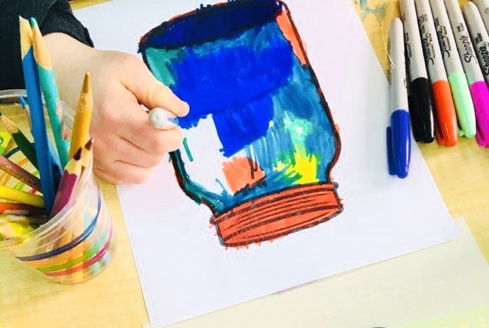 A small hand painting a jar over a table using sharpie colors.