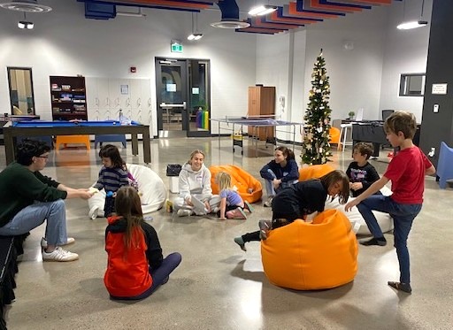A group of children and an adult sit in a circle on bean bags inside a community center decorated with a Christmas tree.