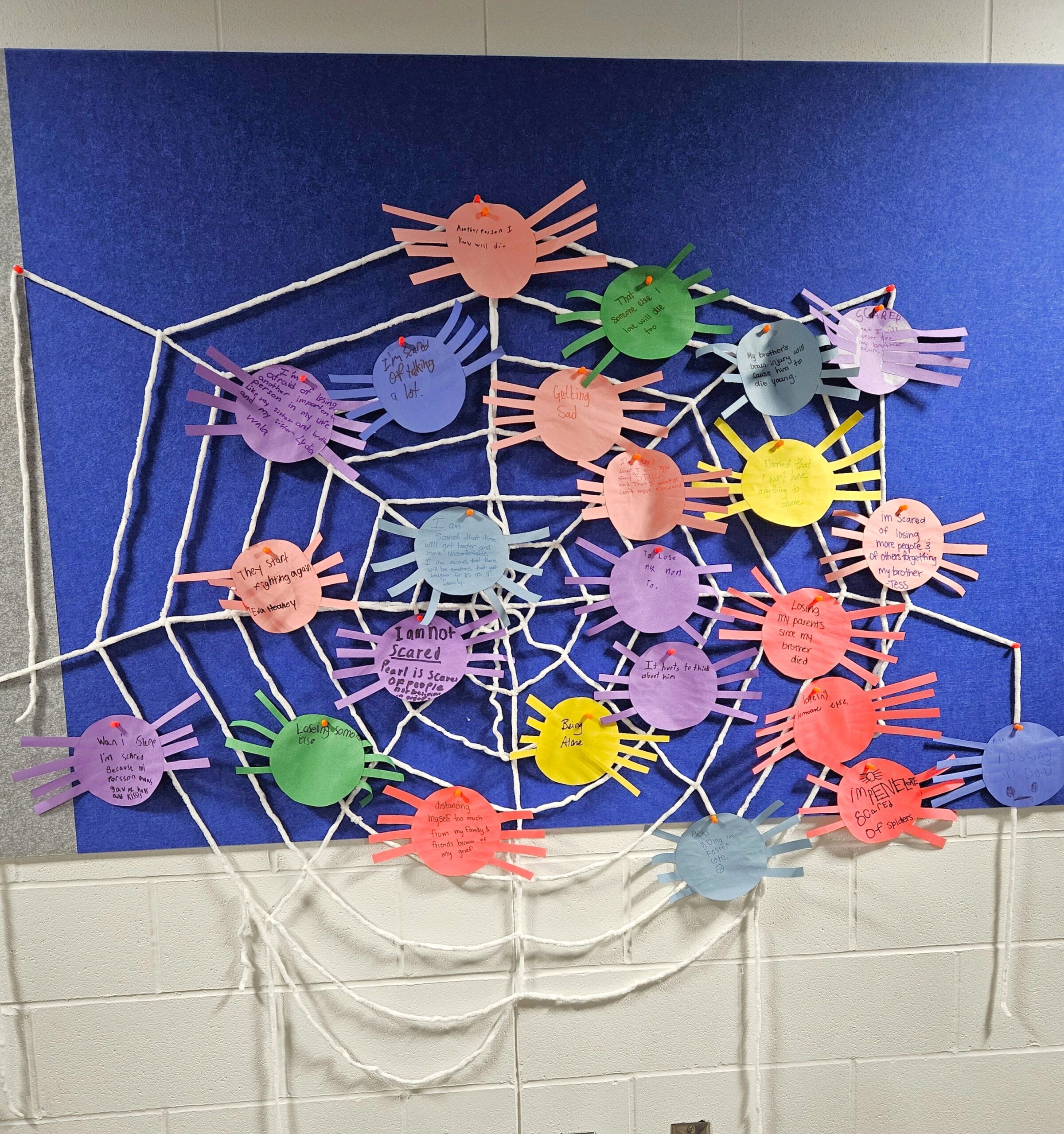 A classroom display of a large spiderweb with colorful paper spiders attached. Each spider has handwritten messages or names on it.