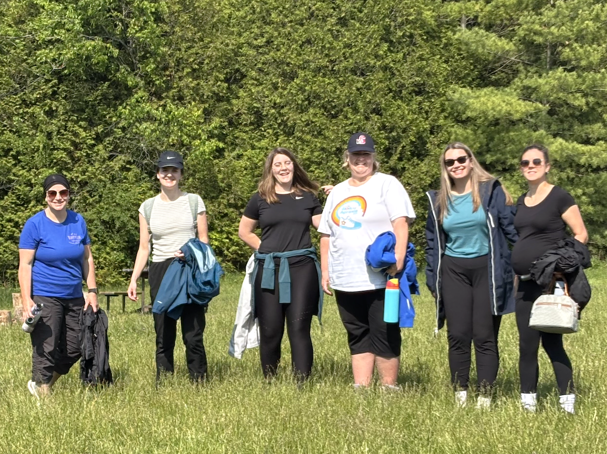 Group of six women standing together outdoors in a grassy field, smiling and dressed for a hiking with trees in the background