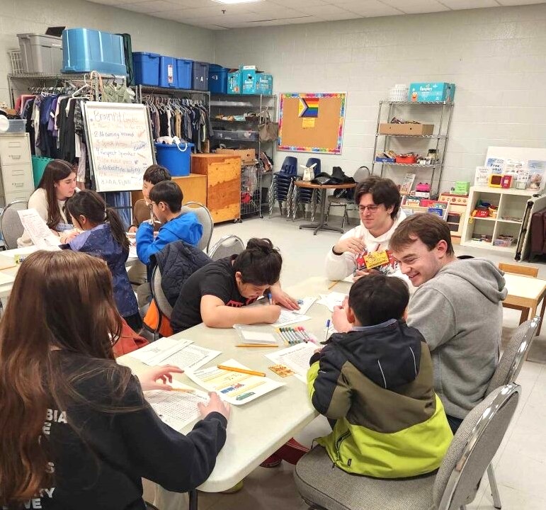 Volunteers and children sitting around tables working on reading and writing activities in a community classroom