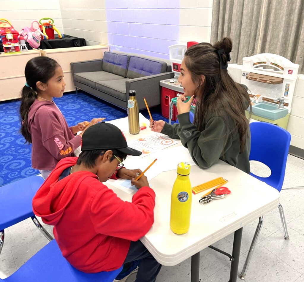 Two children and a volunteer sitting at a small table doing writing activities in a classroom