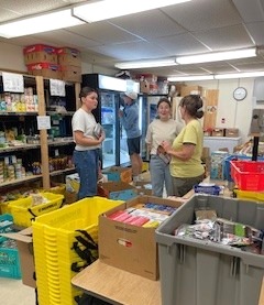 Four people organizing food items inside a food bank, surrounded by bins, boxes, and shelves stocked with supplies.