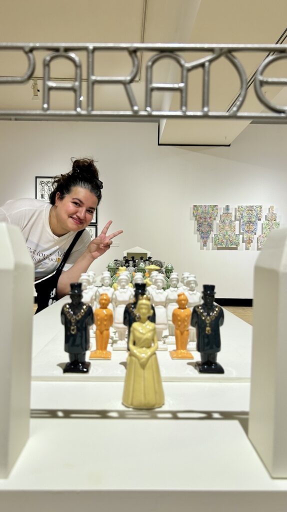 Smiling person making a peace sign while leaning beside an art installation of colorful figurines arranged in rows inside a gallery of Guelph.