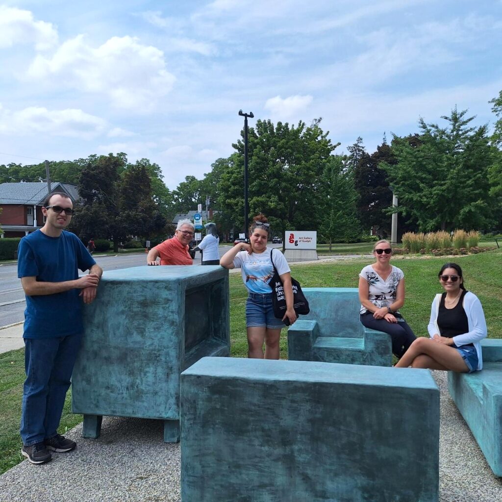 Five people posing outdoors with large blue-green sculptures shaped like furniture, in a grassy park area on a sunny day.