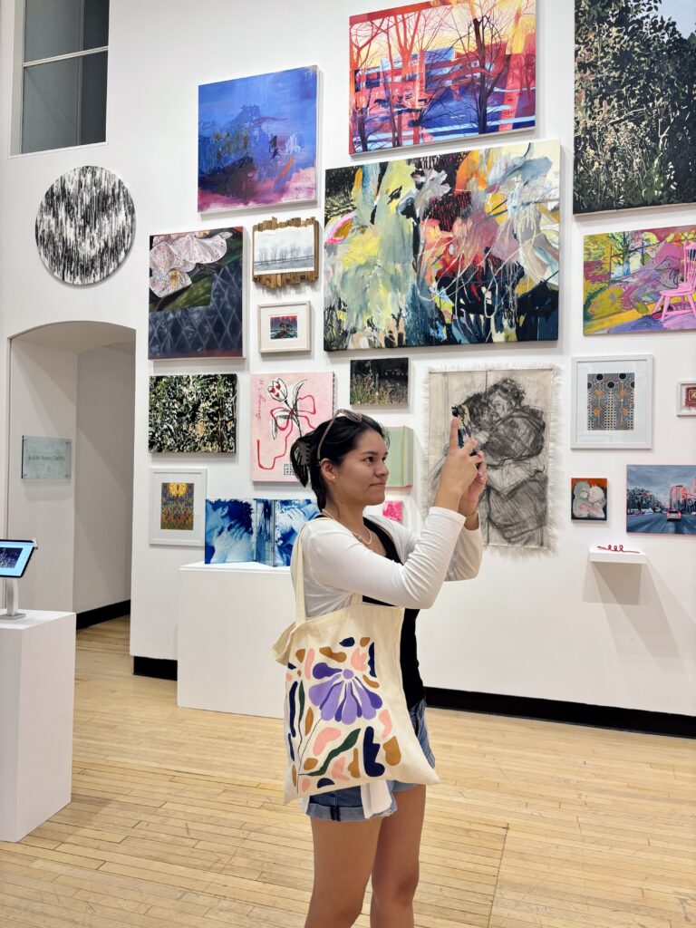 Person standing in an art gallery of Guelph, looking at a large wall filled with colorful paintings and mixed-media artworks of various sizes.