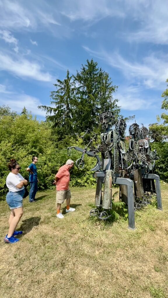Three people outdoors viewing a large metal sculpture made of welded parts, set in a grassy area with trees in the background.