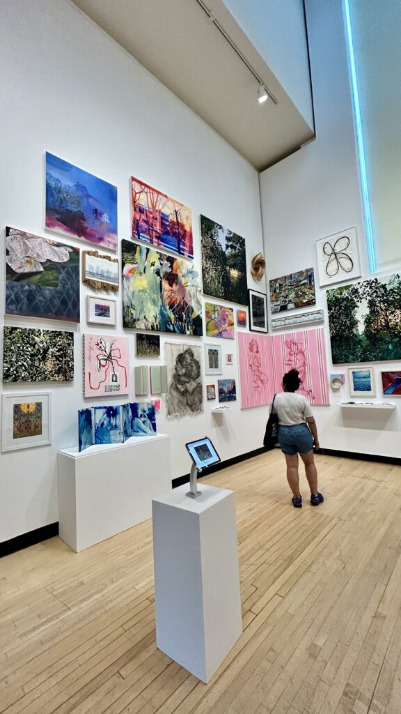 Person standing in an art gallery, looking at a large wall filled with colorful paintings and mixed-media artworks of various sizes.