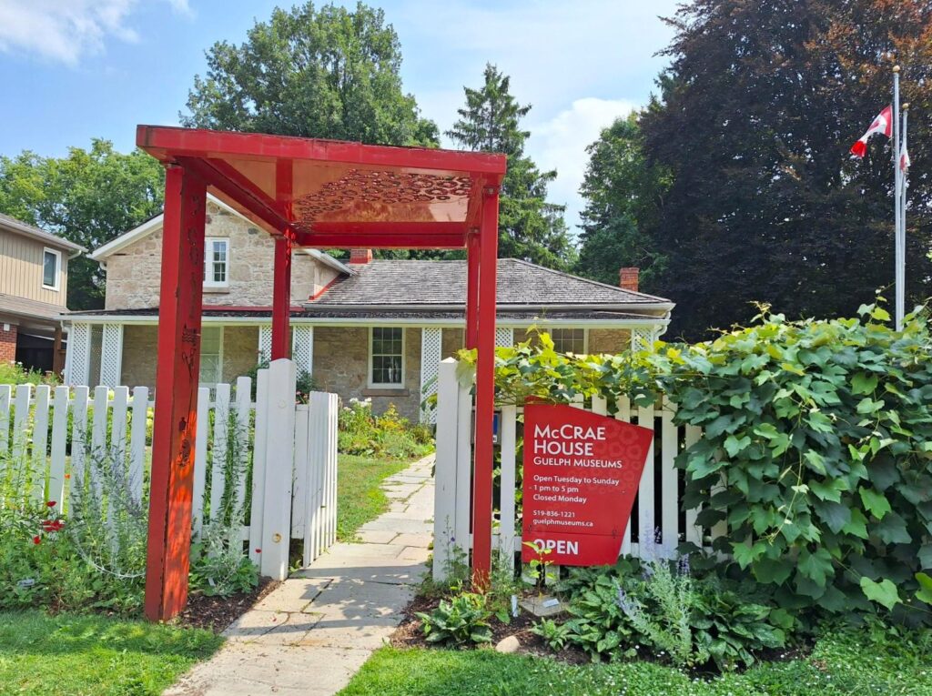 Entrance to McCrae House in Guelph, with a bright red archway, white picket fence, and a sign indicating the museum is open.