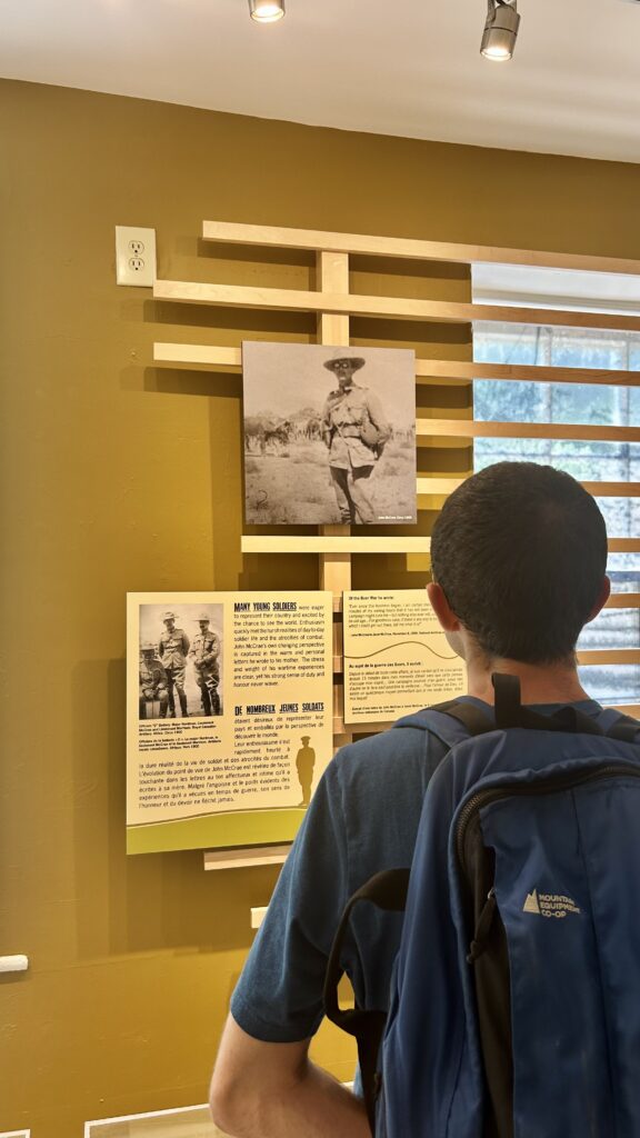 Person with a blue backpack reading an exhibit display about John McCrae, featuring historical photographs and text on a yellow wall.