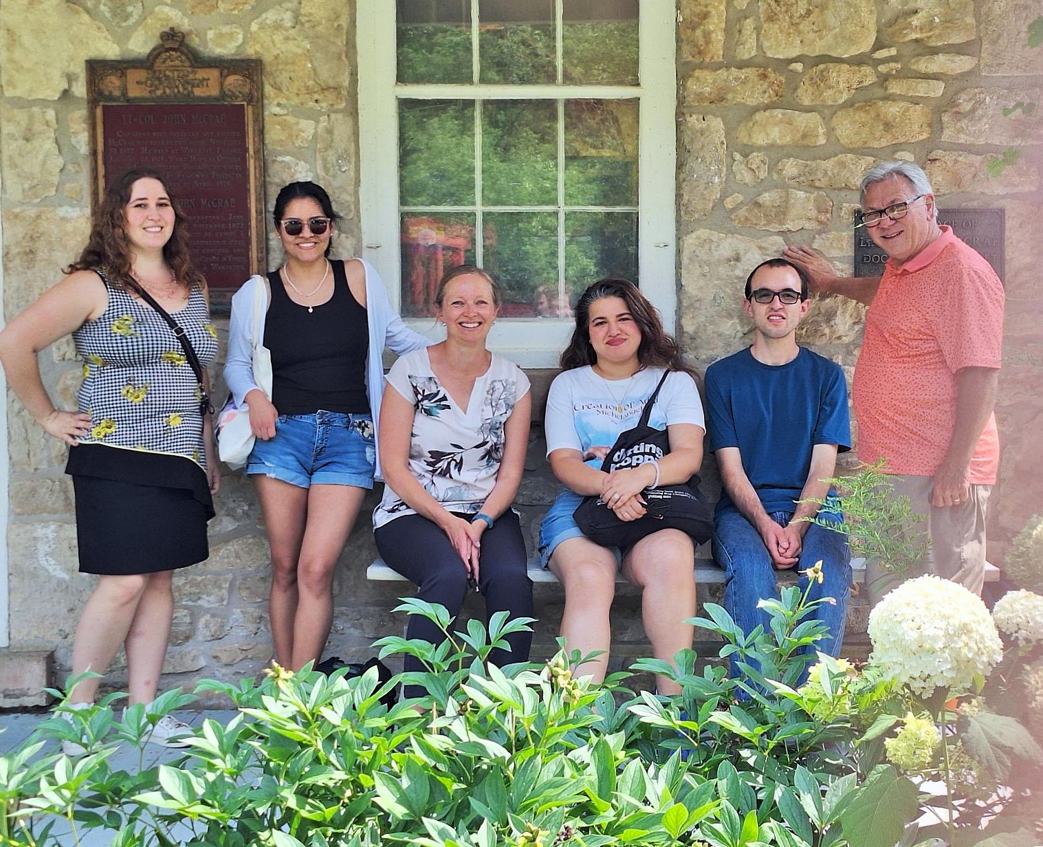 Group of six people smiling in front of a stone building with a window and plaques that belongs to The Mc Crae House, surrounded by greenery on a sunny day.