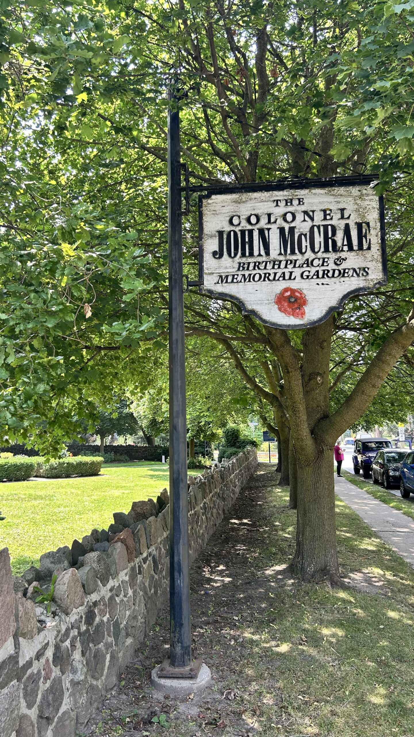 Sign for The Colonel John McCrae Birthplace & Memorial Gardens on an old-time-y sign in front of a tree in the front garden