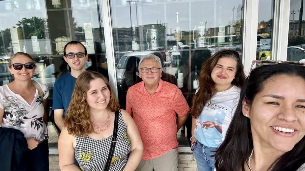 Group of six people standing outside a storefront with glass windows reflecting parked cars and trees.