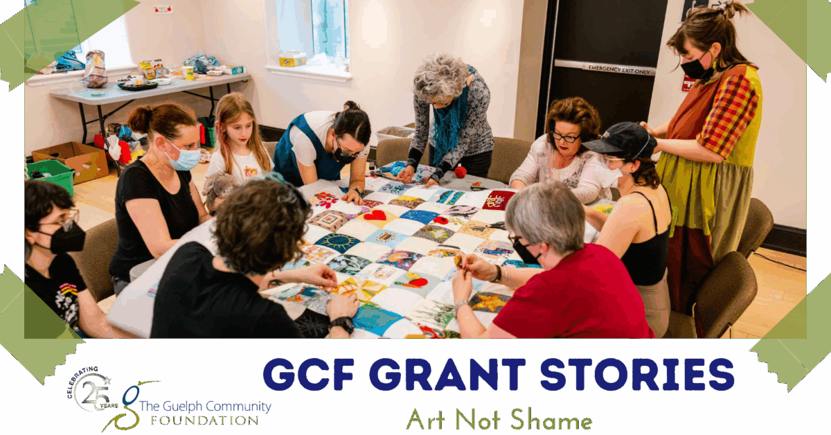 A group of people gathered around a table collaboratively working on a colorful patchwork quilt in a community room.
