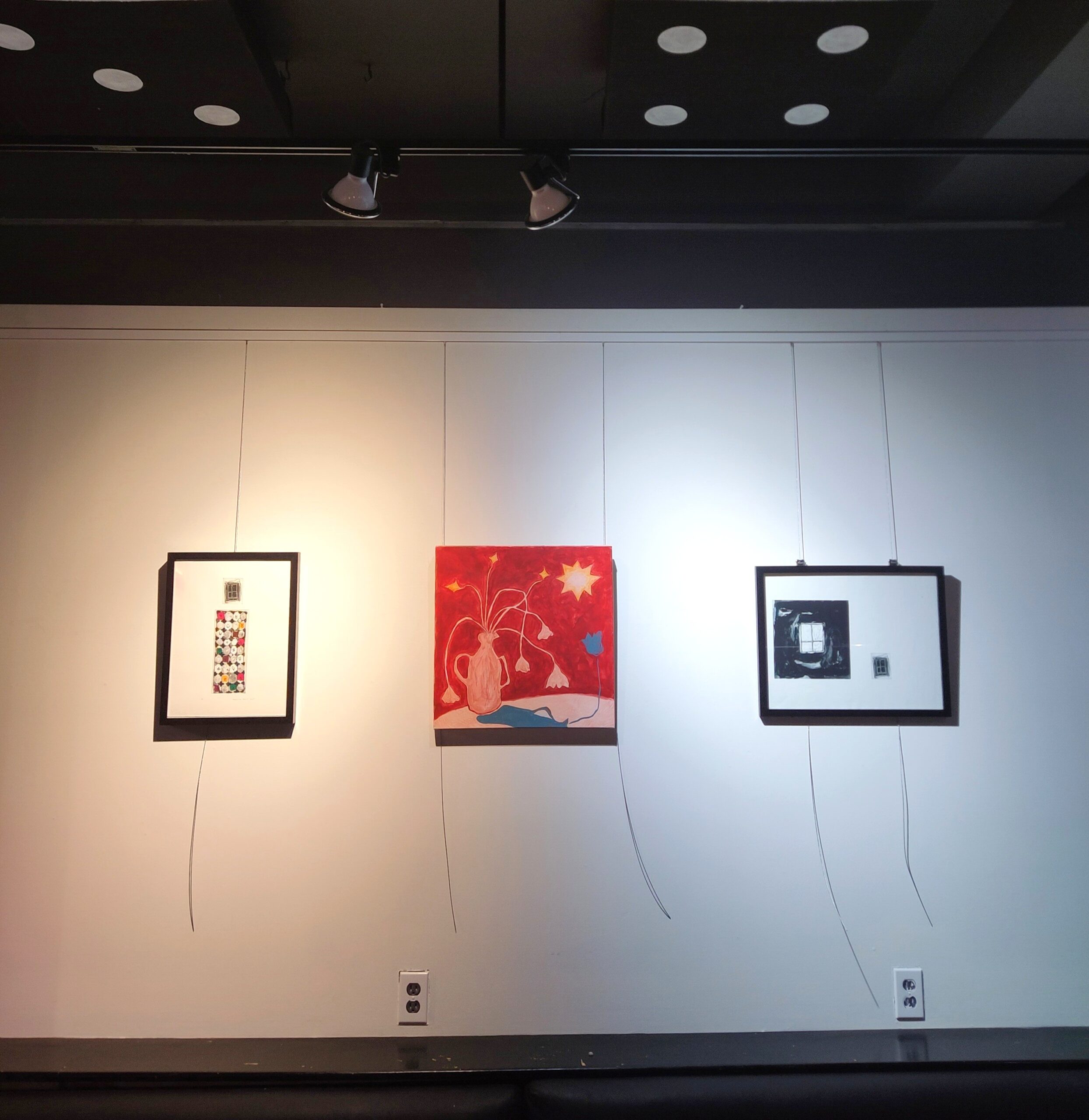 Three framed artworks hang on a white gallery wall under spotlights, featuring abstract and colorful designs with visible hanging wires.