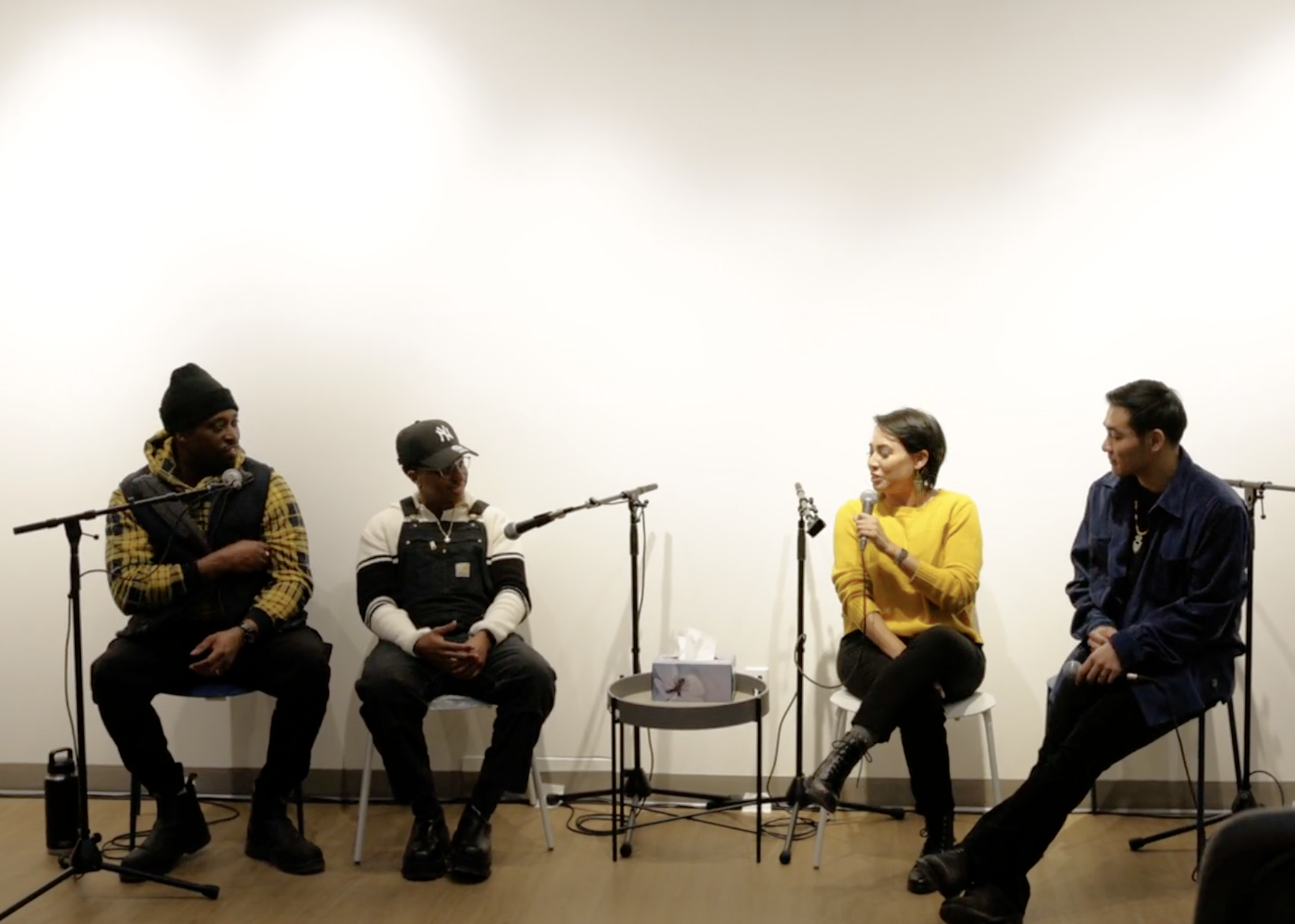Four people seated on chairs in a panel discussion setup with microphones and a small table with tissues between them.