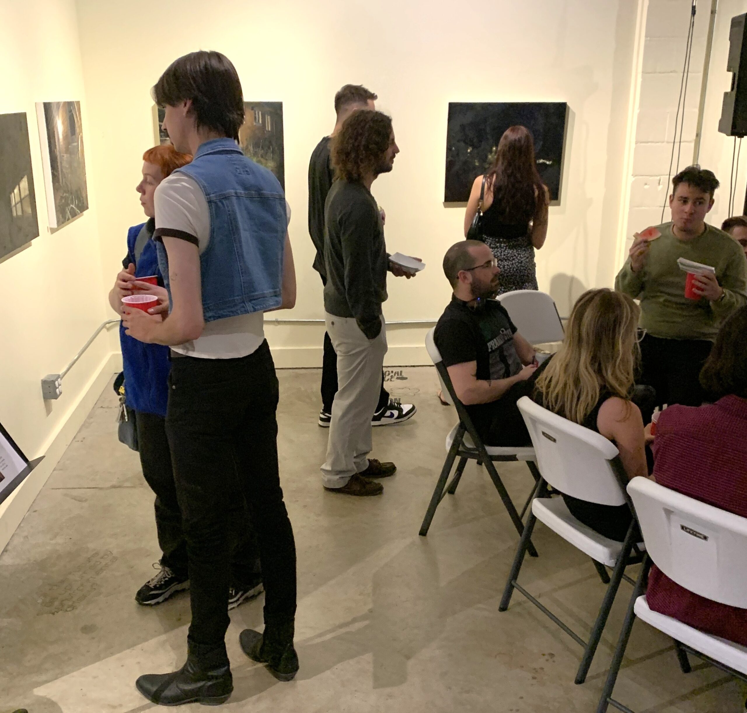 People mingling and viewing artwork in a small gallery space with white walls and concrete floors.