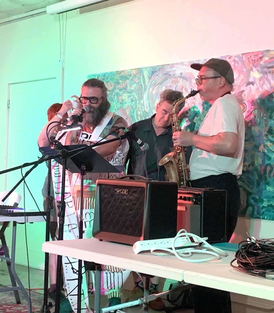 Four musicians performing indoors with microphones, a saxophone, and amplifiers on a table in front of them.