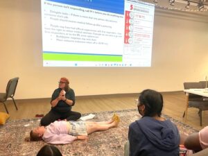 Instructor demonstrates first aid techniques on a person lying on a carpeted floor during a training session in a modern room with string lights.
