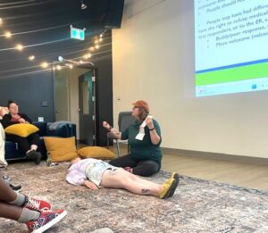 Instructor demonstrates first aid techniques on a person lying on a carpeted floor during a training session in a modern room with string lights.
