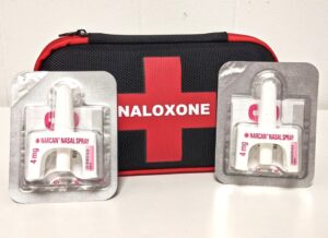 Two Narcan nasal spray 4 mg packages displayed in front of a black and red naloxone emergency kit with a red cross symbol.
