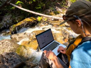 Person wearing a cap and sunglasses working on a laptop while sitting on rocks beside a flowing mountain stream surrounded by greenery.