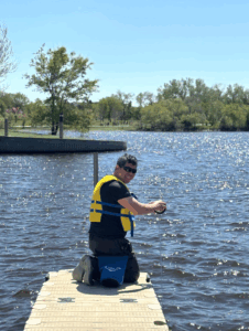 Person wearing a yellow life jacket kneeling on a dock while fishing in a sparkling lake on a sunny day.