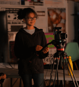 A young woman standing behind professional video cameras on tripods, smiling and adjusting equipment in a studio setting with soft lighting.
