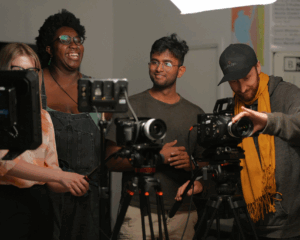 Three people standing behind professional video cameras on tripods, smiling and adjusting equipment in a studio setting with soft lighting.