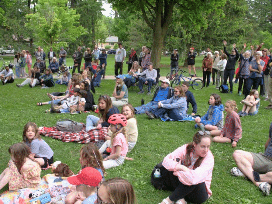 Large group of people, including families and children, seated on blankets and the grass at an outdoor community event in a park, watching a performance or presentation.