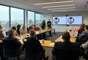 Photo of female presenter at podium with guests at tables watching the presentations in board room