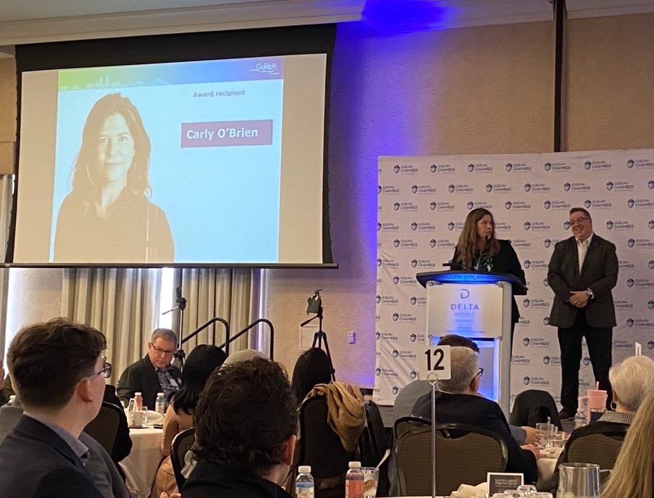 Two speakers, Carly and Cam, delivering a presentation at a formal business event with audience seated at round tables in a banquet hall with the projector screen reading 'Carly O'Brien Award Recipient'