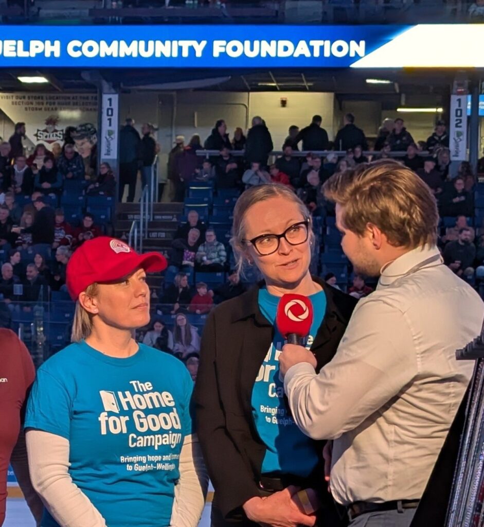 Glenna and Sarah wearing their blue 'The Home for Good Campaign' shirts being interviewed by a reporter holding a red microphone in a crowded indoor hockey arena.