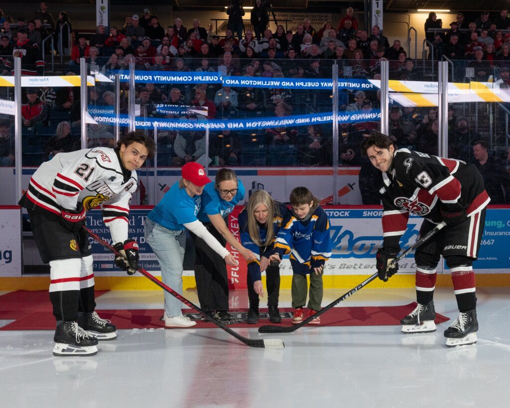 A ceremonial puck drop takes place on the ice rink with Sarah and Glenna and others with the Storm and Attack OHL hockey players with the crowd watching on.