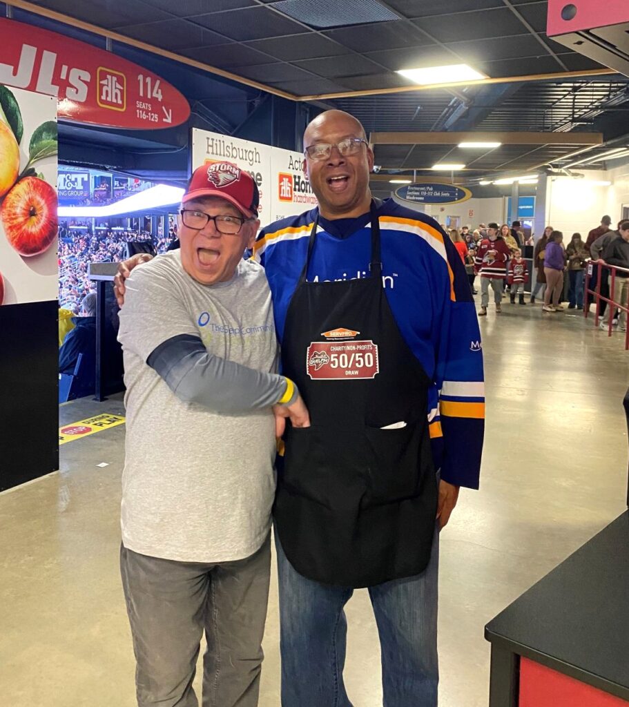 Two men posing together in a stadium concourse, one wearing a blue hockey jersey and black apron with a 50/50 raffle logo, the other in a gray shirt and jeans.