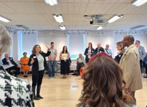 Group of people standing in a circle in a bright room, listening to a woman speaking and gesturing with her hand.