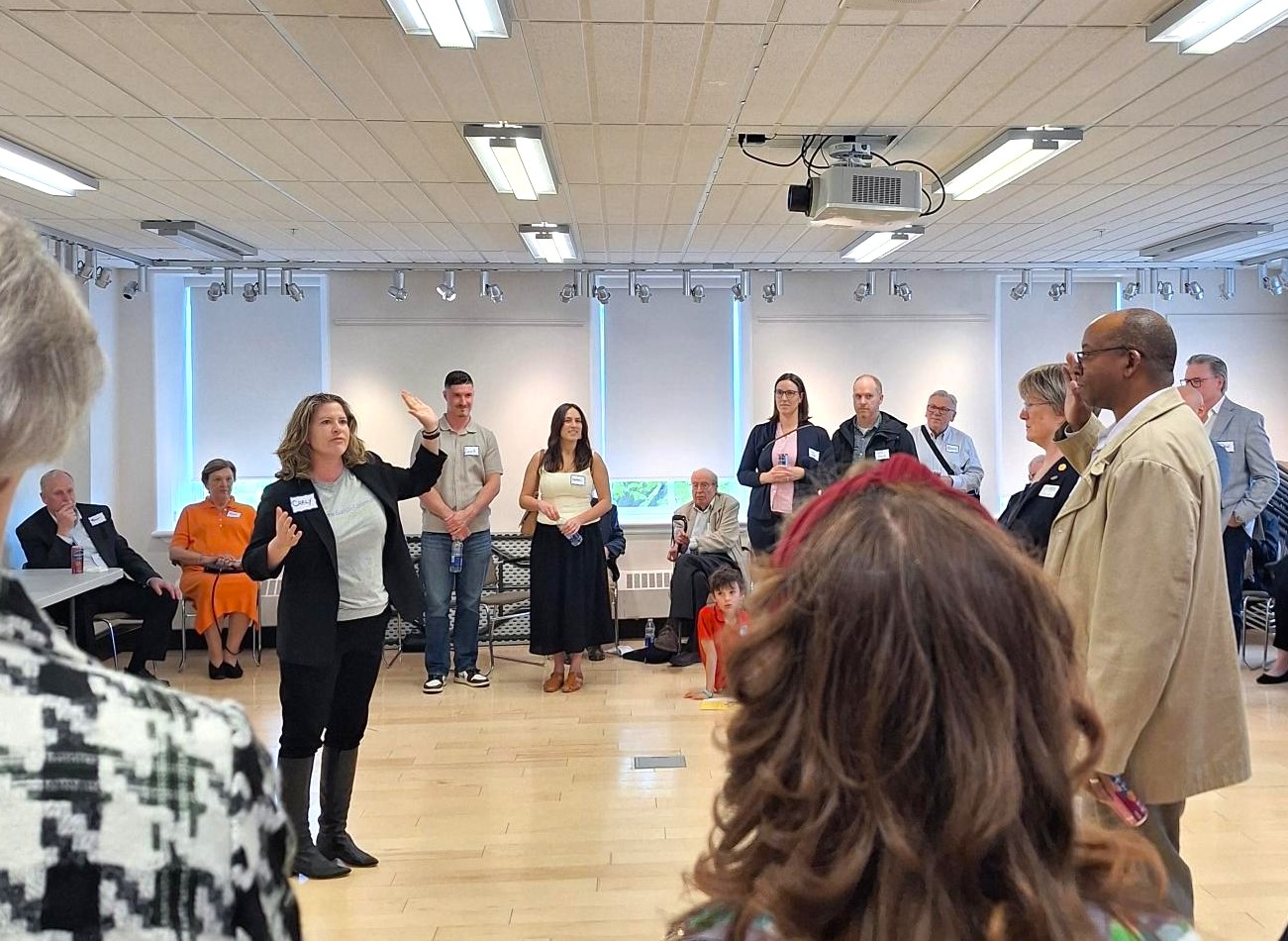 Group of people standing in a circle in a bright room, listening to a woman speaking and gesturing with her hand.