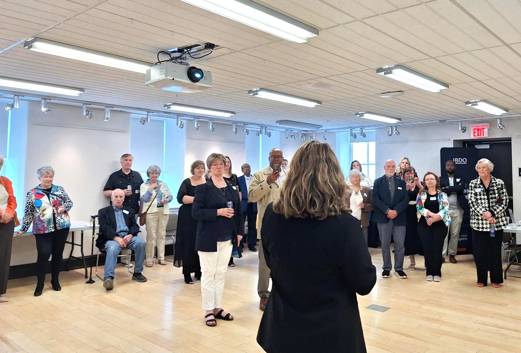 A woman addressing a diverse group of adults gathered in a bright, modern room with wooden floors and ceiling lights.