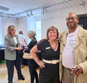 Two people posing together at a social gathering in a bright room with wooden floors and white walls.