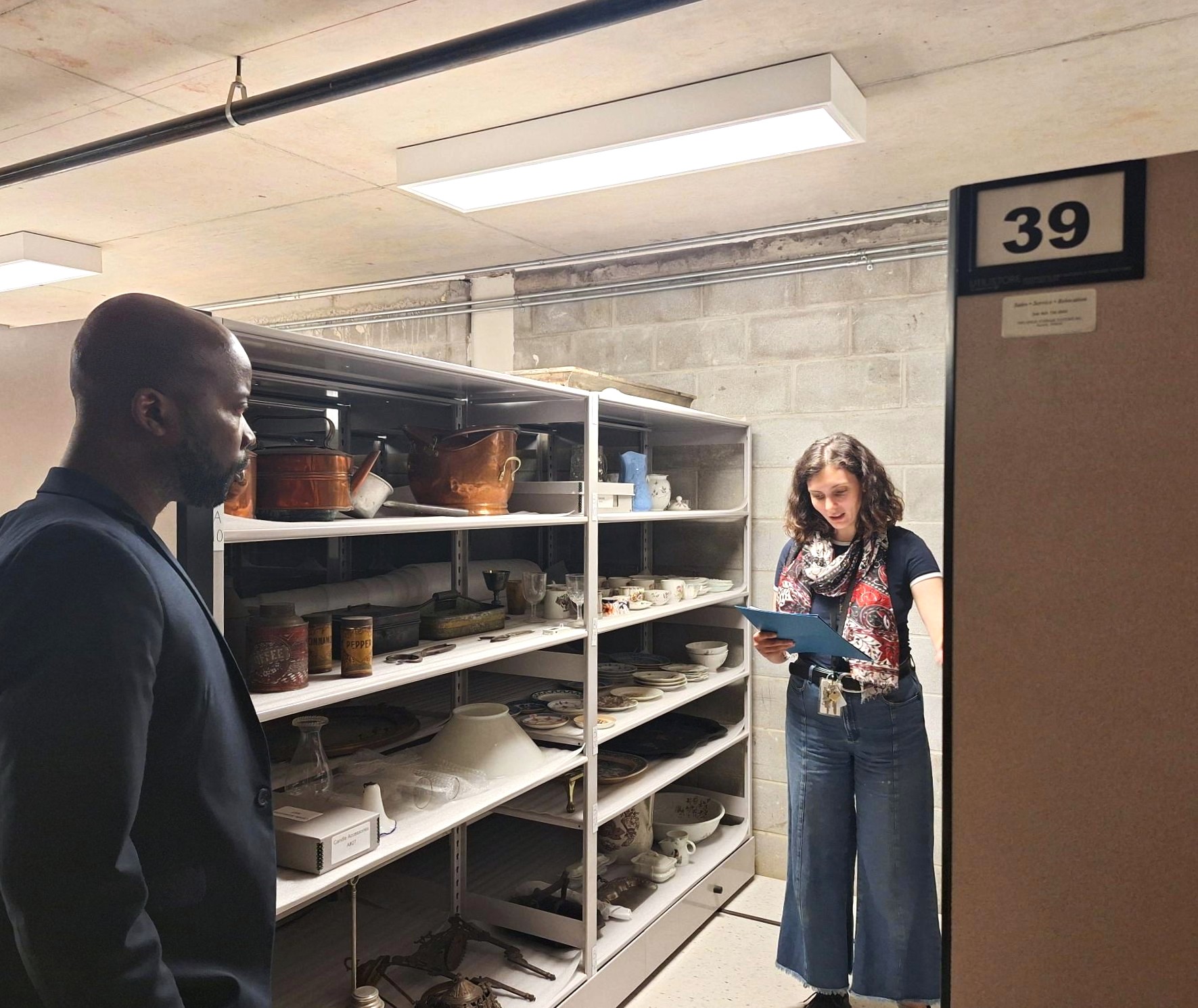 Two people examining archival shelves filled with various pottery and ceramic items in a basement storage room
