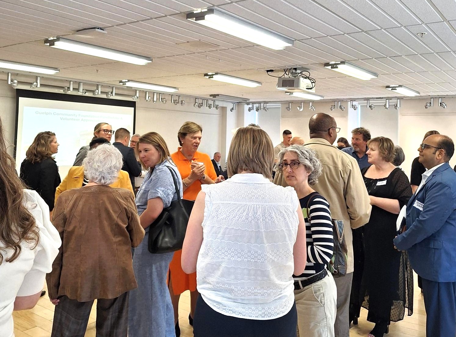 A diverse group of adults engaged in conversation during a networking event in a bright conference room with a projector screen.