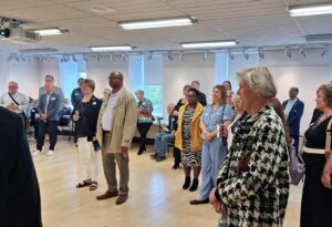 A diverse group of adults standing attentively in a bright, spacious room with wooden floors and large windows.