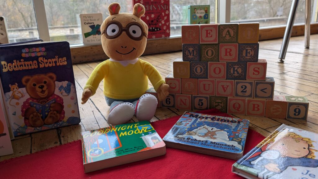 CG 2025 Children's Reading Room (3) A doll in a yellow sweater sits among children's books and wooden alphabet blocks arranged on a wooden floor near a window.