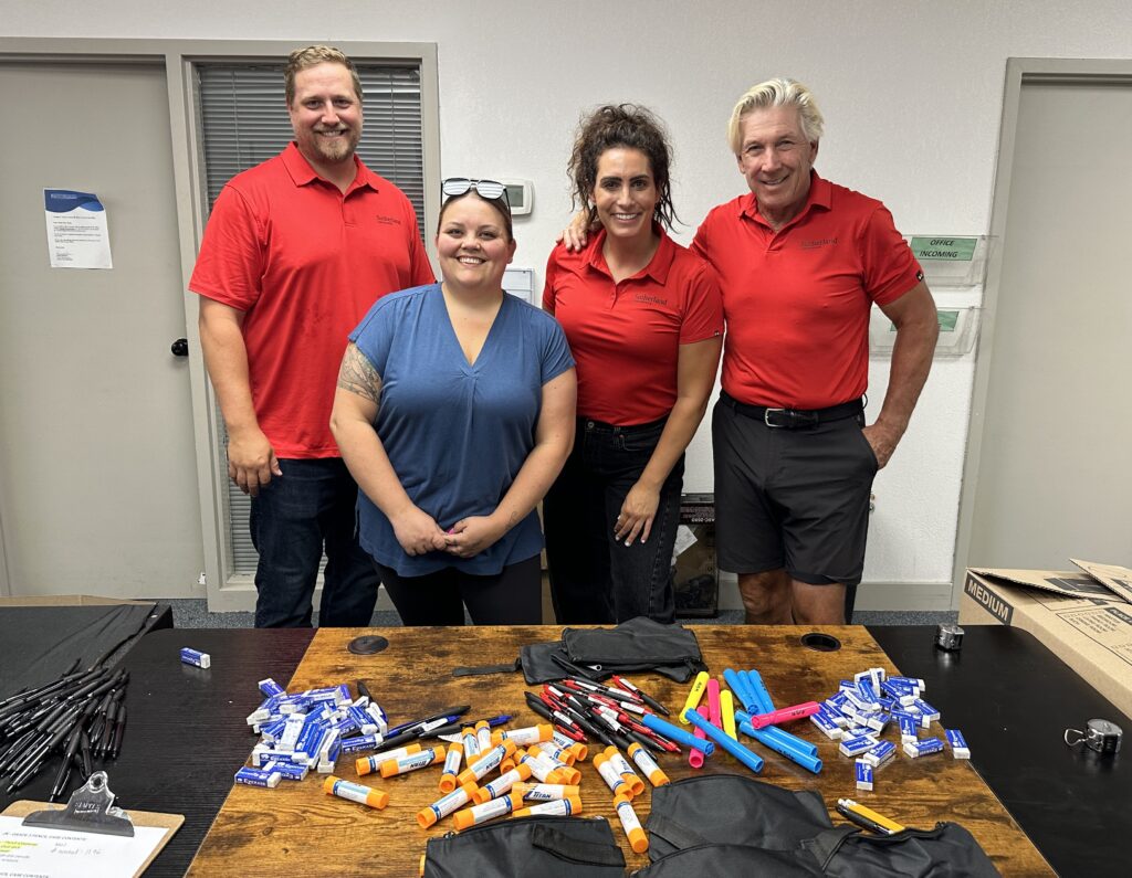 Four people standing behind a table filled with various pens, markers, and small orange containers in an office setting.