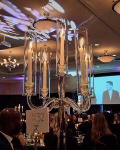 Elegant glass candelabra with lit candles as centerpiece at a formal event with guests seated and a speaker on screen.
