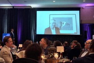 Audience seated at a formal dinner event watching a speaker projected on a large screen on stage.